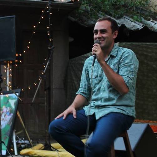 Dan Queirolo sentado num banquinho, cantando ao microfone